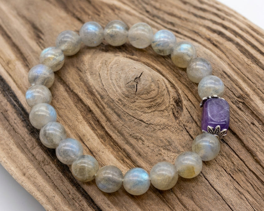 A beaded stretch bracelet made of round light gray-green stone beads with a prominent purple rectangular accent bead, displayed on a piece of natural wood against a white background.