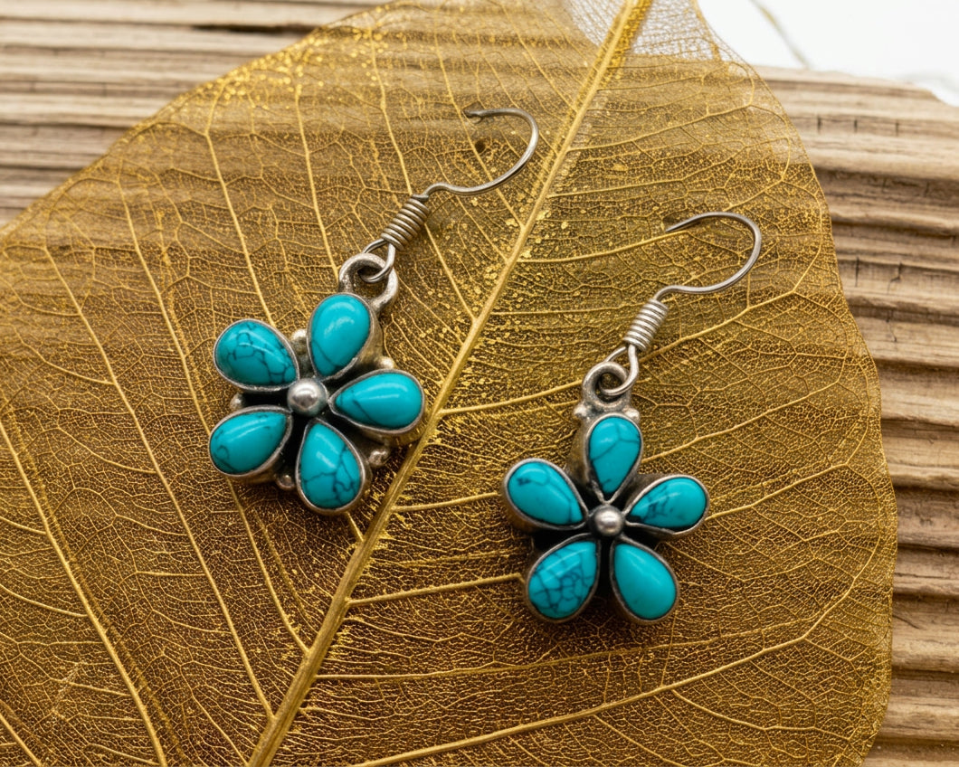 A pair of silver-tone flower dangle earrings with turquoise-blue stone petals, displayed on a gold-toned leaf over a wooden surface.