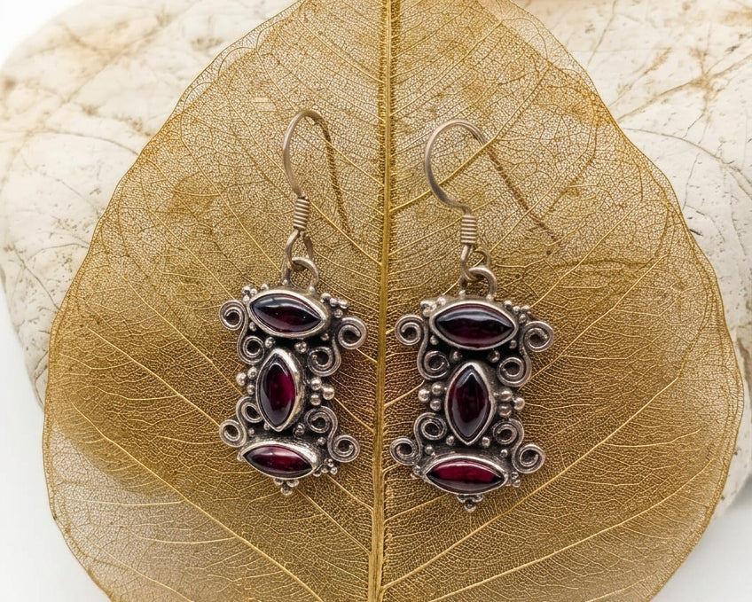 A pair of ornate silver-toned drop earrings with dark red oval and marquise-shaped stones, displayed on a dried brown leaf over a light stone background.