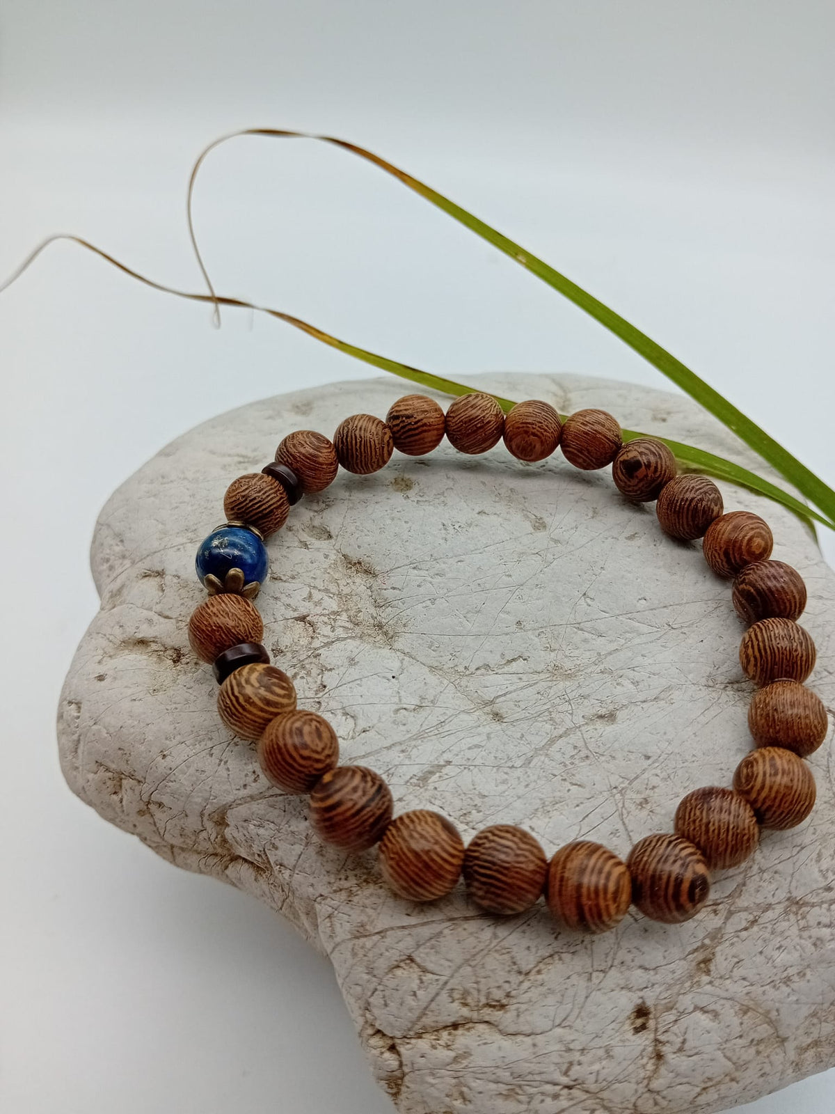 A beaded bracelet made of wenge brown wood beads with a single dark blue lapis lazuli bead and two small dark spacer beads, displayed on a light stone against a white background.