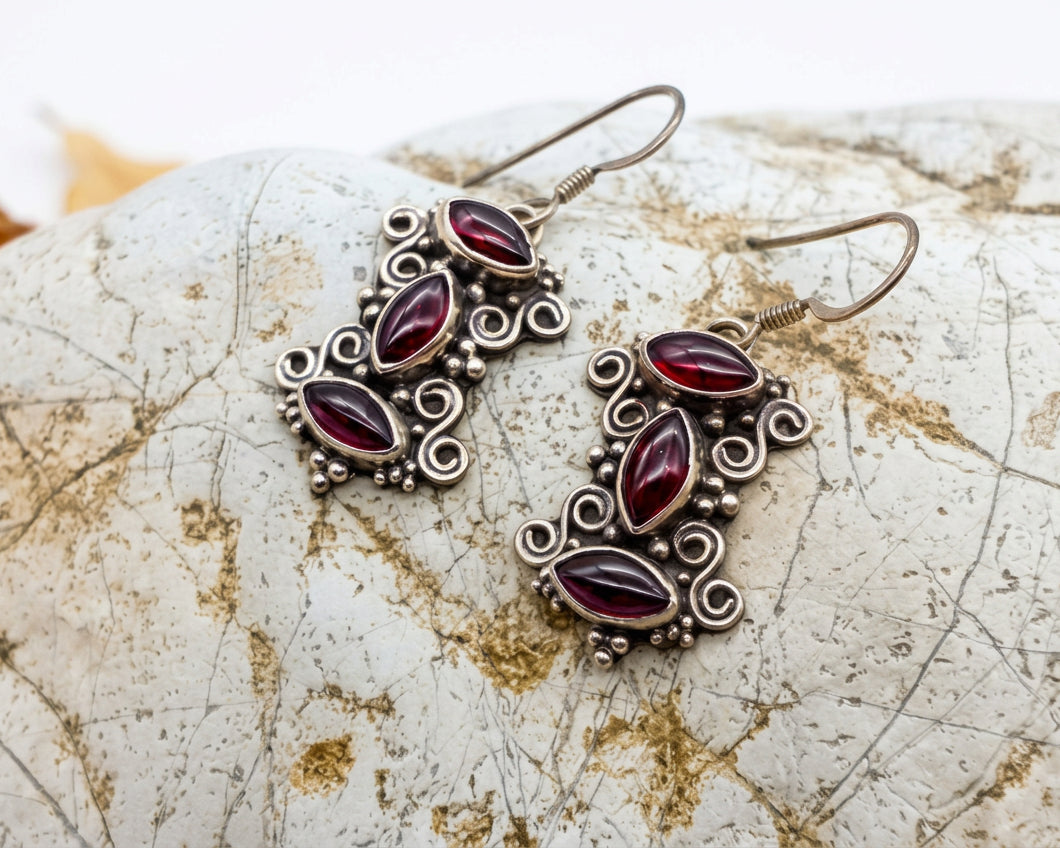A pair of silver-toned drop earrings with marquise-shaped dark red stones and ornate filigree detailing, displayed on a textured light stone prop with dried leaves at the side.