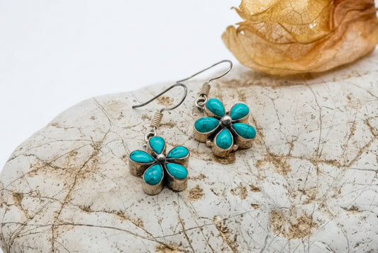 A pair of flower-shaped turquoise drop earrings with silver-toned metal settings and fishhook wires, displayed on a textured beige stone with a dried orange flower prop against a white background.